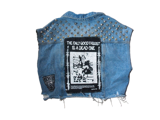 A light blue denim vest laid flat on a tan carpeted floor, featuring silver studs across the shoulders, a large black-and-white graphic patch on the back with the text 'THE ONLY GOOD FASCIST IS A DEAD ONE,' a smaller patch on the left side, and distressed frayed edges at the bottom.