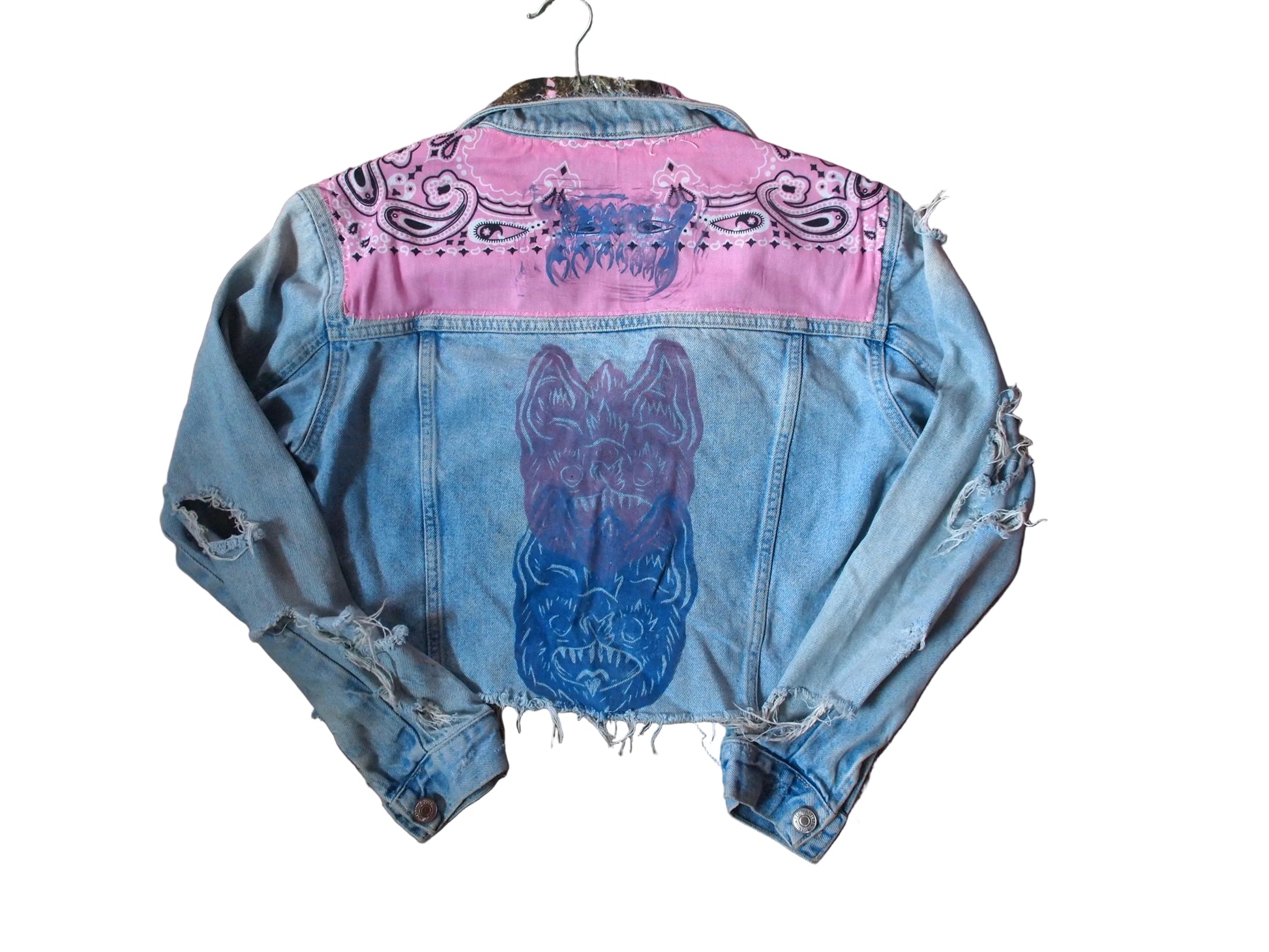 A light blue distressed denim jacket laid flat. The back of the jacket features a bright pink painted section across the shoulders with black paisley-style graphics, and a large blue and purple painted bat face design on the center back.