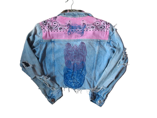 A light blue distressed denim jacket laid flat. The back of the jacket features a bright pink painted section across the shoulders with black paisley-style graphics, and a large blue and purple painted bat face design on the center back.