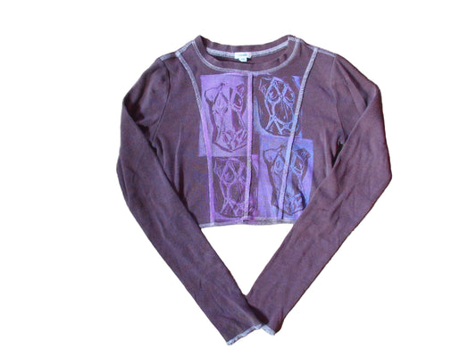 A long-sleeve cropped top in a dusty mauve-purple color, featuring a large lavender graphic print on the front with geometric and abstract patterns, laid flat on a beige carpet.